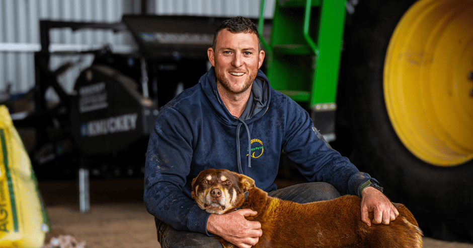 A farmer smiling at the camera as he pas his farm dog whilst crouching in front of a machinery shed.