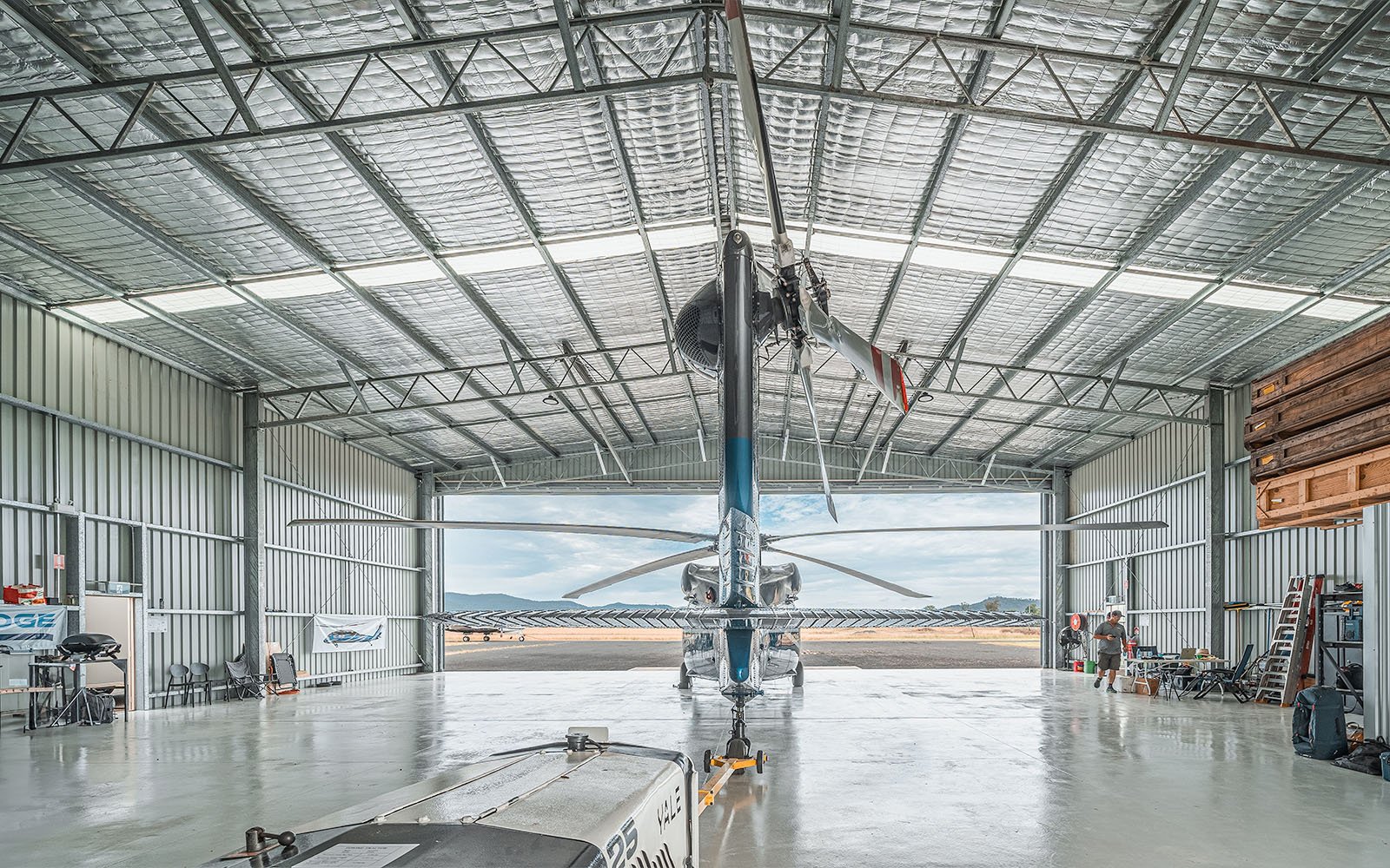 A Black Hawk helicopter seen from behind in a galvanised steel aircraft hangar, with work equipment and seating stores either side, and pallets stacked on top of a built-in office.
